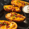 ['Four loaded baked potatoes with cheese, bacon, and chives on a black tray with a side of sour cream.']