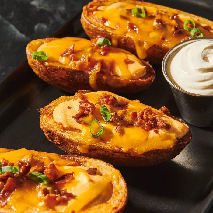['Four loaded baked potatoes with cheese, bacon, and chives on a black tray with a side of sour cream.']