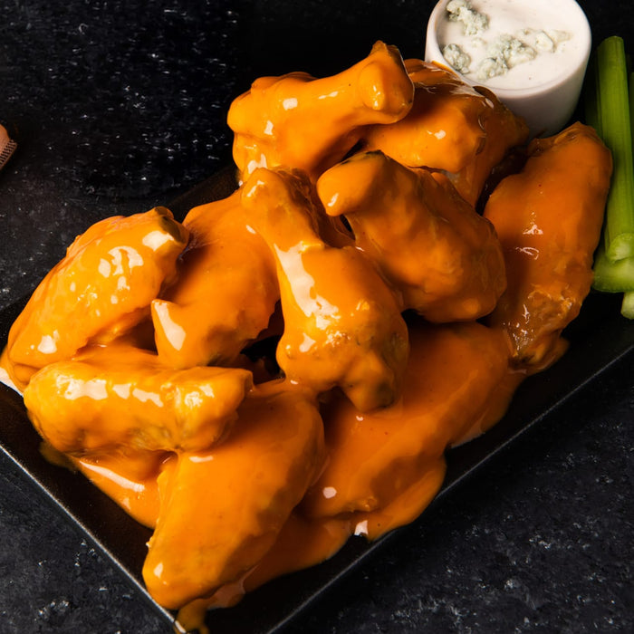A platter of crispy chicken wings is generously coated in Franks Red Hot Regular Wing Hot Sauce Mild 12 Oz and served with a side of blue cheese dressing and fresh celery sticks.