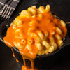 A small black cast iron pot filled with creamy macaroni and cheese is topped with a generous pour of Franks Red Hot Regular Wing Hot Sauce Mild 12 Oz dripping down the sides.