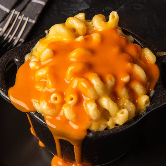 A small black cast iron pot filled with creamy macaroni and cheese is topped with a generous pour of Franks Red Hot Regular Wing Hot Sauce Mild 12 Oz dripping down the sides.