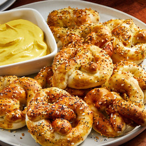 A white bowl filled with Frenchs Yellow Regular Mustard 12 Oz sits next to a plate of soft pretzels topped with everything bagel seasoning.