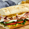 A hearty deli sandwich on a long baguette is generously layered with sliced turkey, ham, cheese, fresh vegetables, and Frenchs Yellow Regular Mustard 12 Oz.