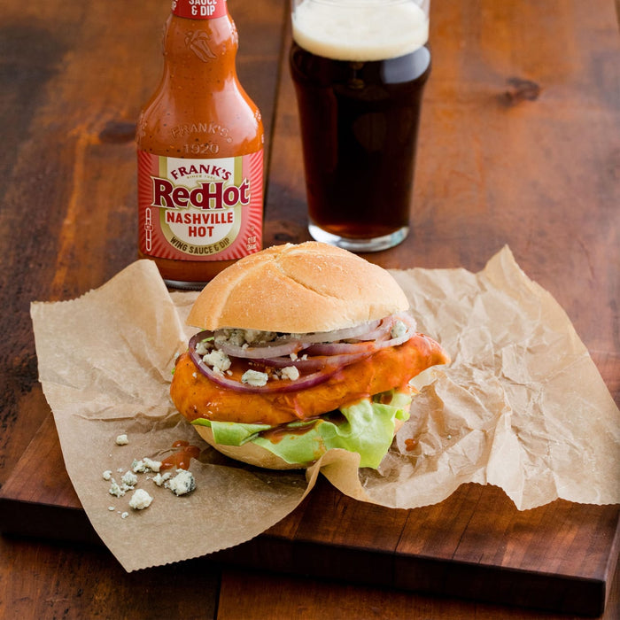 A buffalo chicken sandwich topped with red onions and blue cheese crumbles sits on parchment paper next to a bottle of Franks Red Hot Regular Wing Hot Sauce Hot 12 Oz and a glass of dark beer.