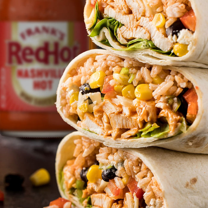 A stack of three chicken burritos filled with rice, corn, and black beans sits in front of a bottle of Franks Red Hot Regular Wing Hot Sauce Hot 12 Oz.