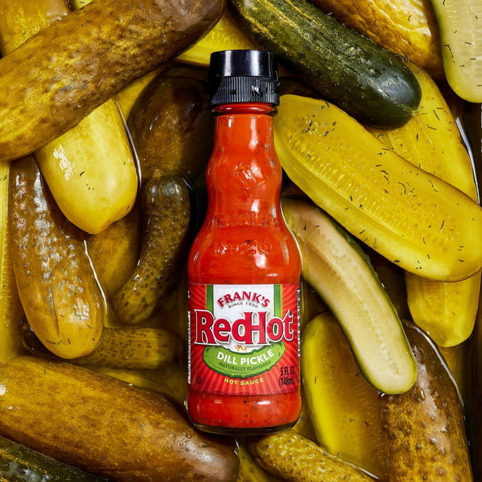 ["A bottle of Frank's RedHot Dill Pickle hot sauce sits among a pile of dill pickles."]