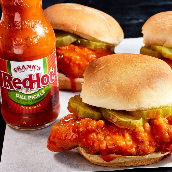 ["A bottle of Frank's RedHot Dill Pickle hot sauce next to a chicken sandwich with pickles."]