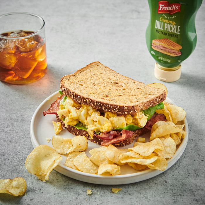 ["A sandwich with egg salad, bacon, and lettuce on a plate with potato chips next to a glass of iced tea and a bottle of French's Creamy Dill Pickle Mustard Spread."]