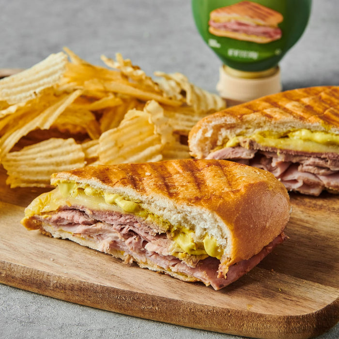 ['A Cuban sandwich with mustard and ham on a wooden cutting board with potato chips.']