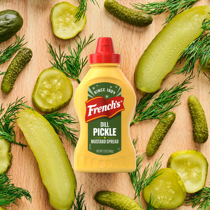 A bottle of Frenchs Dill Pickle Sandwich Spread Regular Inverted Squeeze Bottle Rp 12 Oz is artfully arranged on a wooden cutting board, surrounded by fresh dill, whole gherkins, and sliced dill pickles.