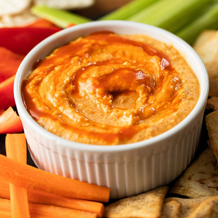 A bowl of vibrant orange hummus drizzled with a swirl of sauce is surrounded by colorful vegetables and pita chips, creating an inviting appetizer spread.