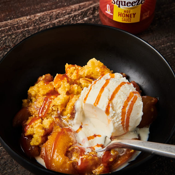 ['A bowl of peach cobbler topped with vanilla ice cream and a drizzle of Squeeze Hot Honey Sauce.']