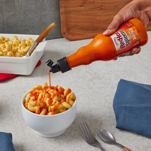 ["A hand is squeezing Frank's RedHot Squeeze Creamy Buffalo Sauce onto a bowl of macaroni and cheese."]