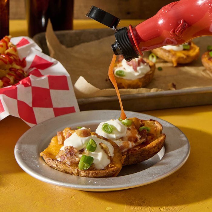 A bottle of hot sauce is being poured onto potato skins topped with cheese, bacon, and sour cream, with a basket of fries in the background.