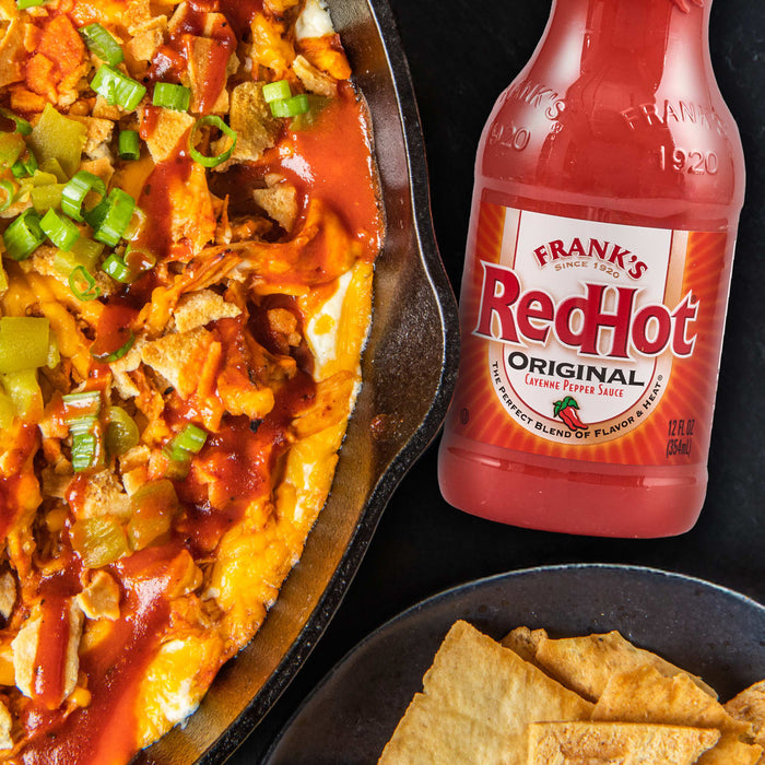 A bottle of Frank's RedHot Original hot sauce sits next to a skillet of loaded nachos topped with cheese, green onions, and hot sauce.