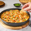 A skillet filled with buffalo chicken dip is shown with a hand dipping a cracker into it, garnished with blue cheese and green onions.