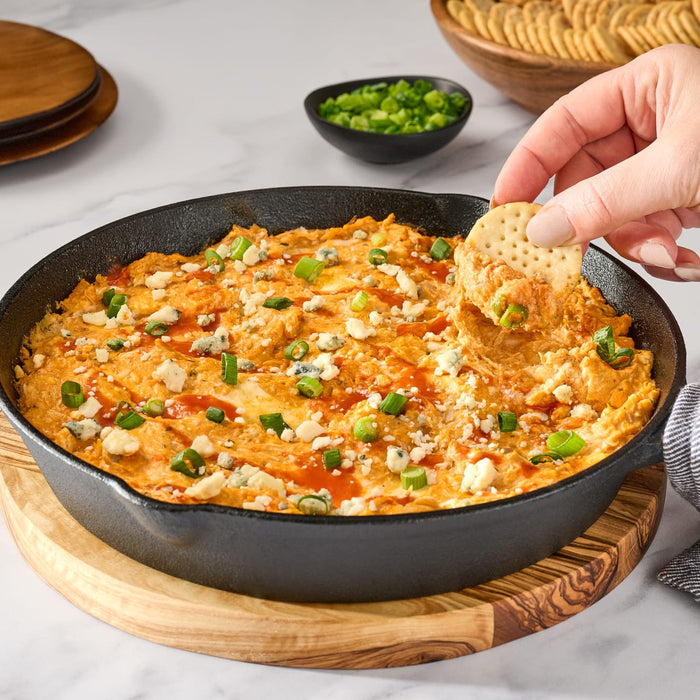 A skillet filled with buffalo chicken dip is shown with a hand dipping a cracker into it, garnished with blue cheese and green onions.