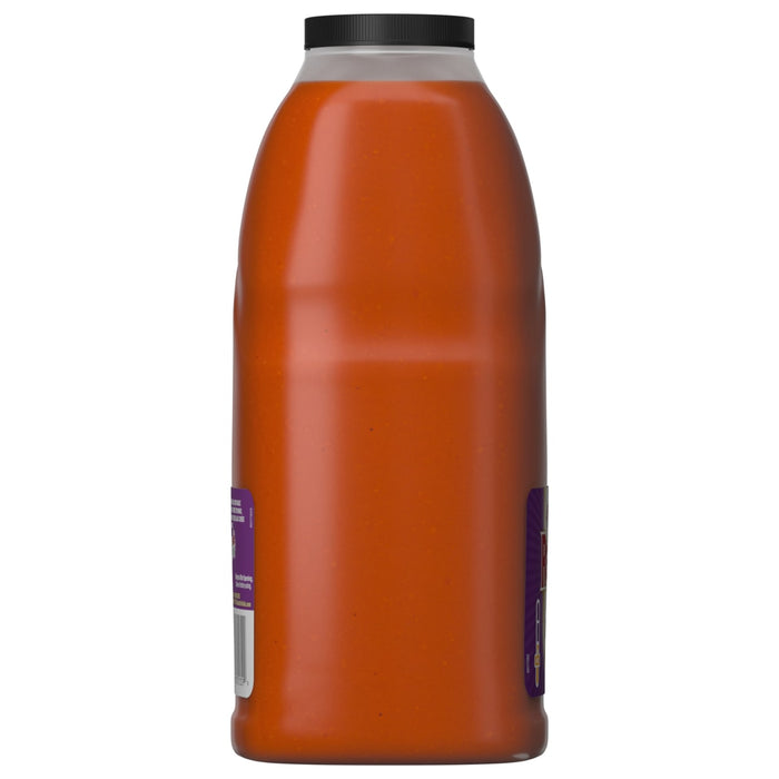 A side view of a large, clear plastic bottle of Franks Red Hot Garlic Buffalo Wing Hot Sauce Regular 64 Oz shows the thick, orange-red sauce within, sealed by a black cap.