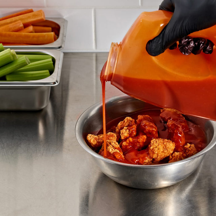 A person wearing a black glove pours Franks Red Hot Mango Habanero Wing Hot Sauce Regular 64 Oz from a large container over a bowl of crispy fried chicken wings next to fresh celery and carrots.