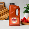 A large jug of Franks Red Hot Mango Habanero Wing Hot Sauce Regular 64 Oz sits on a white countertop next to fresh habanero peppers and a mango.