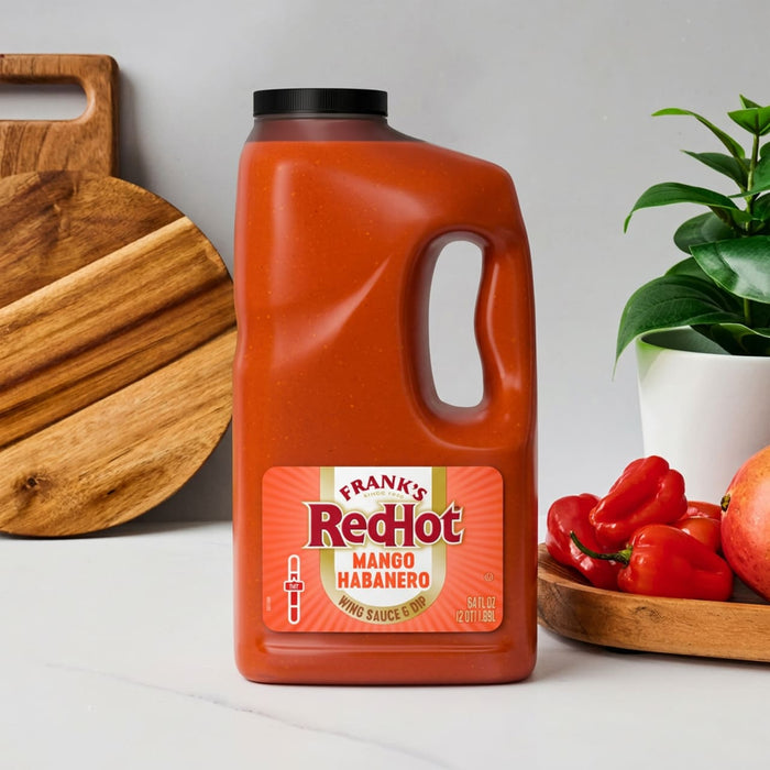 A large jug of Franks Red Hot Mango Habanero Wing Hot Sauce Regular 64 Oz sits on a white countertop next to fresh habanero peppers and a mango.