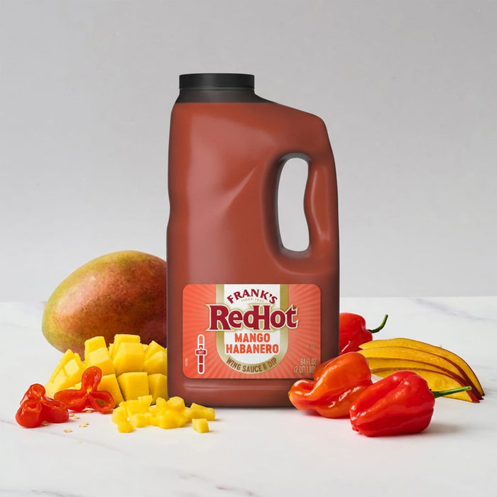 A large jug of Franks Red Hot Mango Habanero Wing Hot Sauce Regular 64 Oz sits on a marble surface surrounded by a whole mango, diced mango pieces, and fresh habanero peppers.