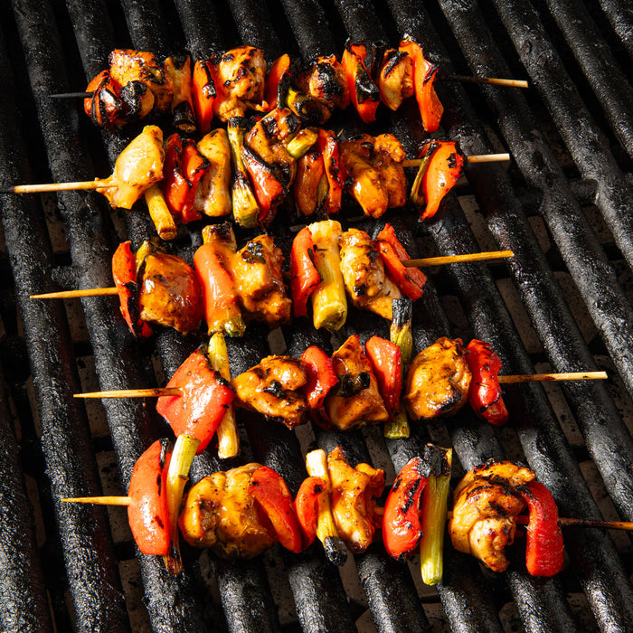 Five skewers of grilled chicken and red bell peppers are glazed with Franks Red Hot Mango Habanero Wing Hot Sauce Regular 64 Oz while cooking on a black grill grate.
