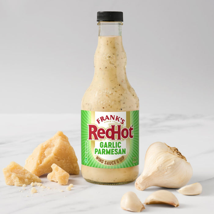 A bottle of Frank's RedHot Garlic Parmesan sauce is displayed with parmesan cheese and garlic cloves.
