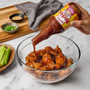 A hand pours Frank's RedHot Wings Sauce General Tao over a bowl of chicken wings.