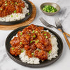 A plate of sesame chicken and rice is shown with a bowl of chopped green onions and two forks.