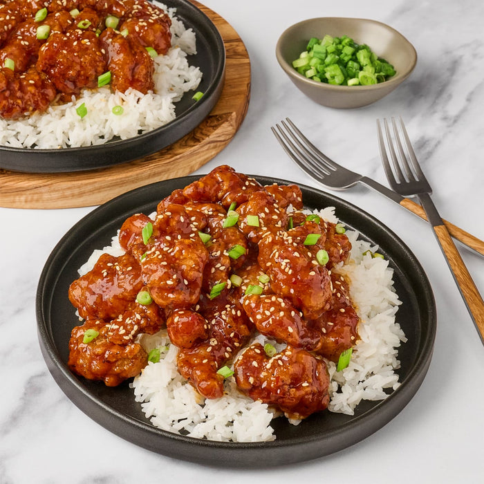 A plate of sesame chicken and rice is shown with a bowl of chopped green onions and two forks.