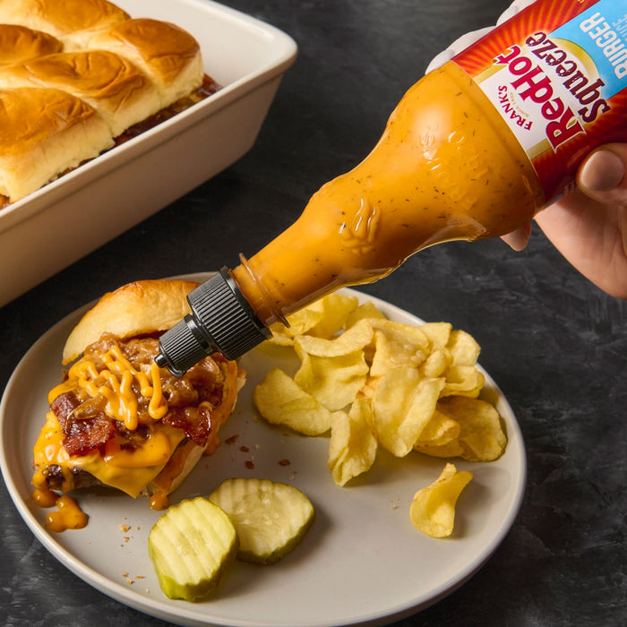 A hand is squeezing Franks RedHot Sauce onto a slider with bacon cheese and caramelized onions.