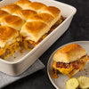 A white baking dish filled with slider buns topped with melted cheese bacon and caramelized onions with one slider on a plate with pickles.