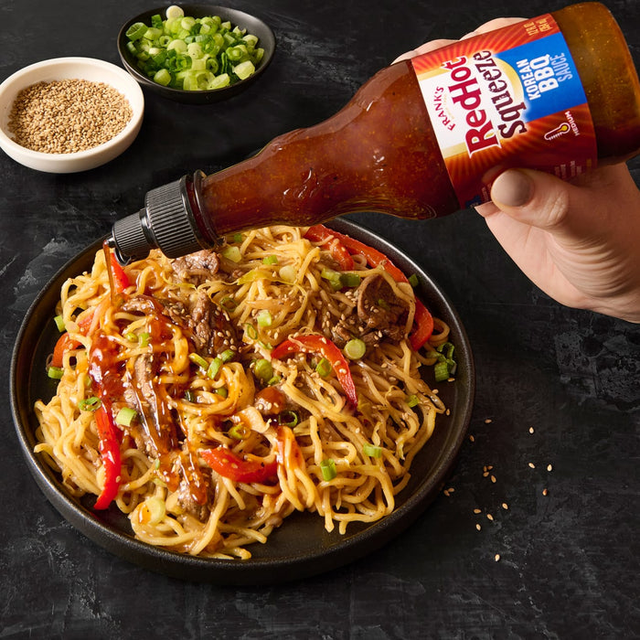 A hand is pouring Franks RedHot Squeeze Korean BBQ Sauce over a bowl of noodles.