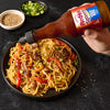 A hand is pouring Franks RedHot Squeeze Korean BBQ Sauce over a bowl of noodles.