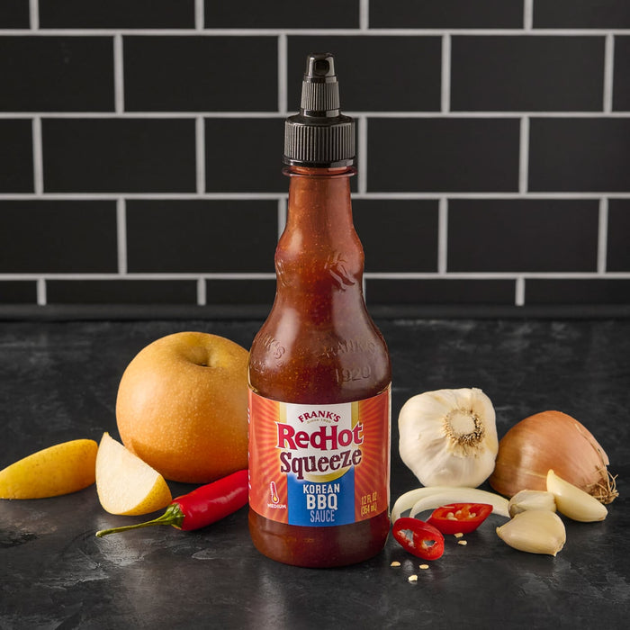 A bottle of Franks RedHot Squeeze Korean BBQ Sauce with some fresh ingredients like garlic onion and chili peppers.