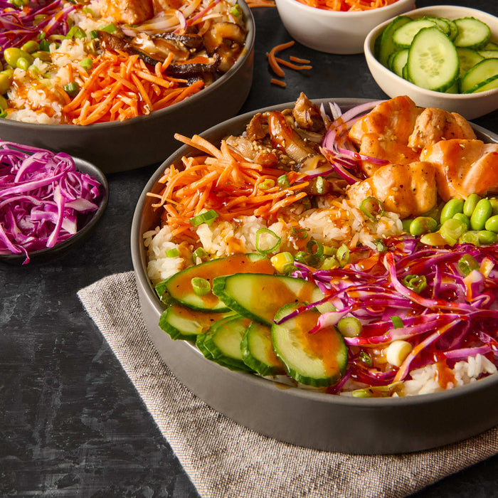 A bowl of rice with salmon carrots cucumbers red cabbage and edamame.