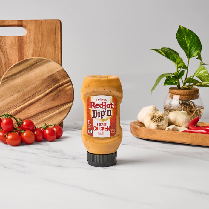 A bottle of Franks RedHot Dipn Secret Chicken Sauce is displayed on a marble surface with tomatoes garlic peppers and wooden cutting boards in the background.
