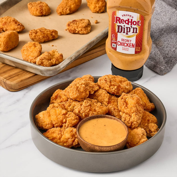 A bowl of fried chicken bites with a small bowl of Franks RedHot Dipn Secret Chicken Sauce in the center and a bottle of the same sauce in the background.