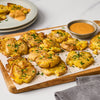 A wooden board is topped with smashed potatoes a bowl of dipping sauce and a gray striped towel with a plate of more potatoes and sauce in the background.