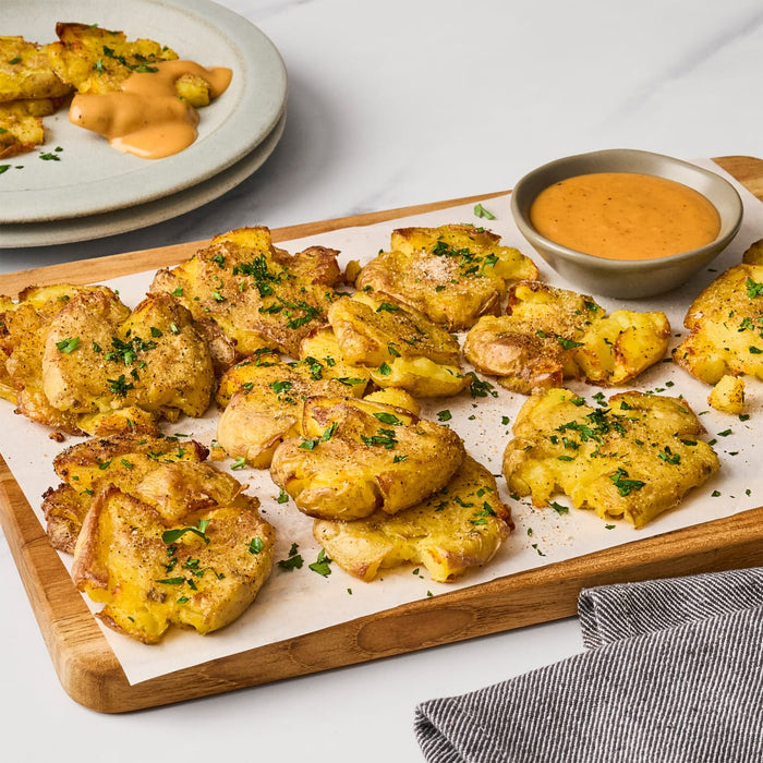 A wooden board is topped with smashed potatoes a bowl of dipping sauce and a gray striped towel with a plate of more potatoes and sauce in the background.