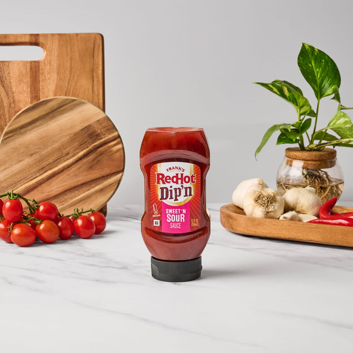 A bottle of Franks RedHot Dipn Sweet N Sour Sauce is centered on a marble surface surrounded by tomatoes garlic peppers wooden boards and a plant.