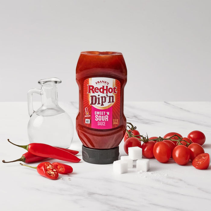 A bottle of Franks RedHot Dipn Sweet N Sour Sauce is displayed with red chili peppers a small carafe of clear liquid sugar cubes and a vine of cherry tomatoes.