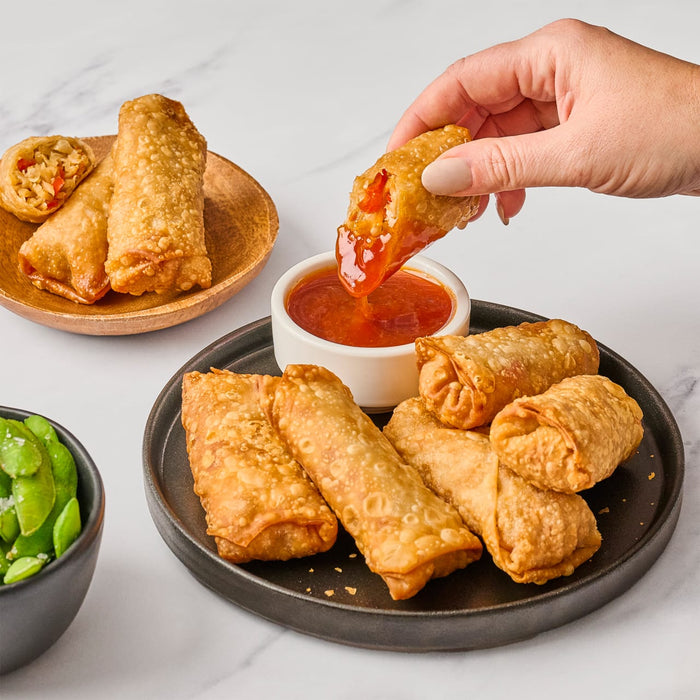 A hand dips a crispy egg roll into a bowl of sweet chili sauce surrounded by more egg rolls on a black plate and a side of edamame.