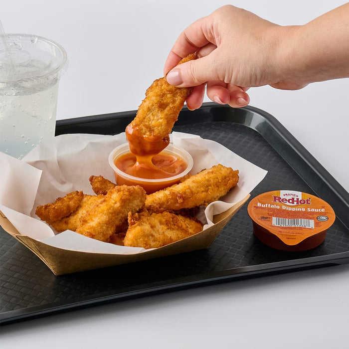 A hand dips a Raising Cane's chicken finger into a sauce, with more chicken fingers and another sauce container on a tray.