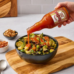 Hand pouring Red Hot sauce into a bowl of salad on a kitchen counter.