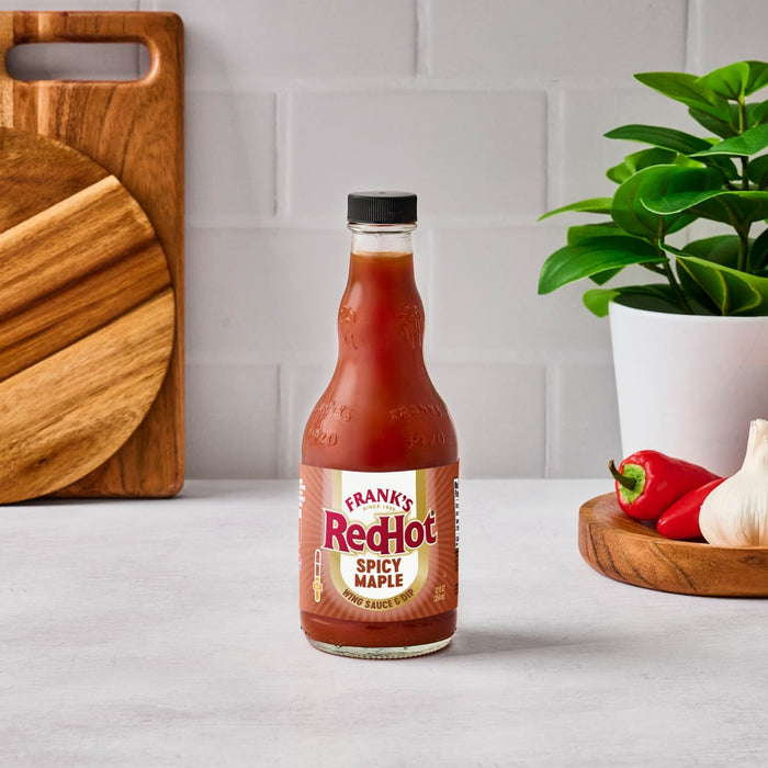 A bottle of Frank's RedHot sauce sits on a counter with garlic and peppers, showcasing the product in a kitchen setting.