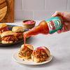 A hand pours Red Duck Foods hot sauce onto a plate of pulled pork sliders topped with pickled onions.