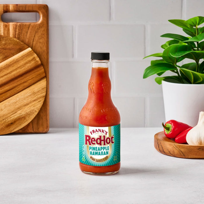 A bottle of Frank's RedHot Pineapple Habanero Sauce sits on a counter with peppers and garlic.