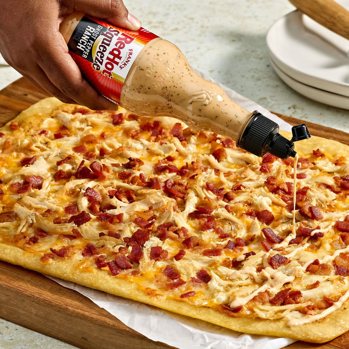 A rectangular pizza topped with chicken and bacon is being drizzled with Ken's Steak House Boom Boom Sauce.